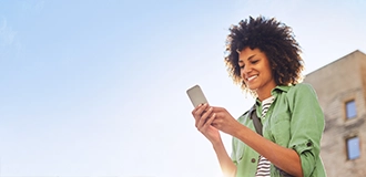 Frau in Freizeitkleidung lächelt auf ihr Handy; Gebäudeteil und blauer Himmel im Hintergrund.