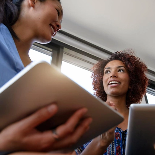 Zwei Frauen sprechen vor einem Fenster und halten Unterlagen.