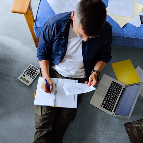 Man sitting in front of a sofa writing in a notebook; laptop, calculator, and documents next to him.