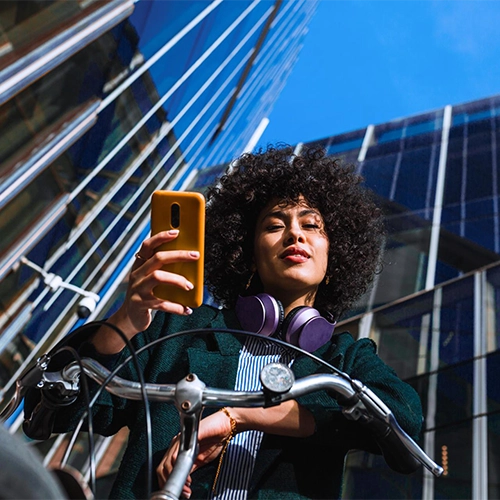 Woman sitting on a bicycle in casual clothes, holding an orange smartphone; buildings and blue sky in the background.